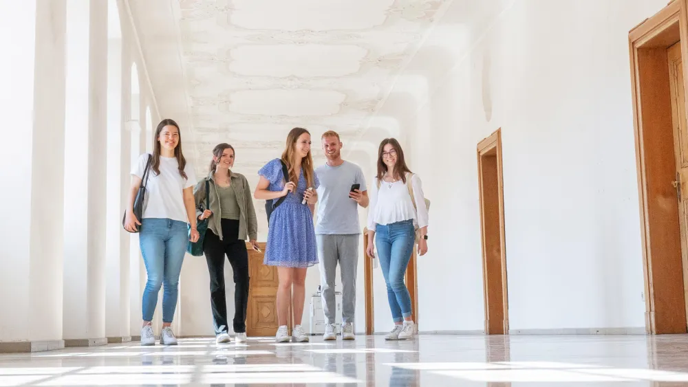 Fünf junge Studierende gehen lächelnd durch einen hellen Korridor mit Büchern und Rucksäcken.