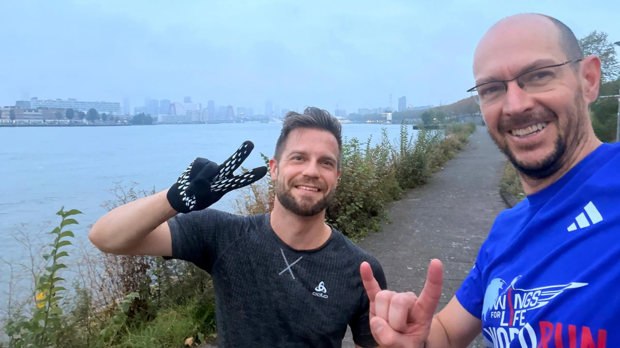 Two smiling men taking a selfie by a lake; one is giving a thumbs-up
